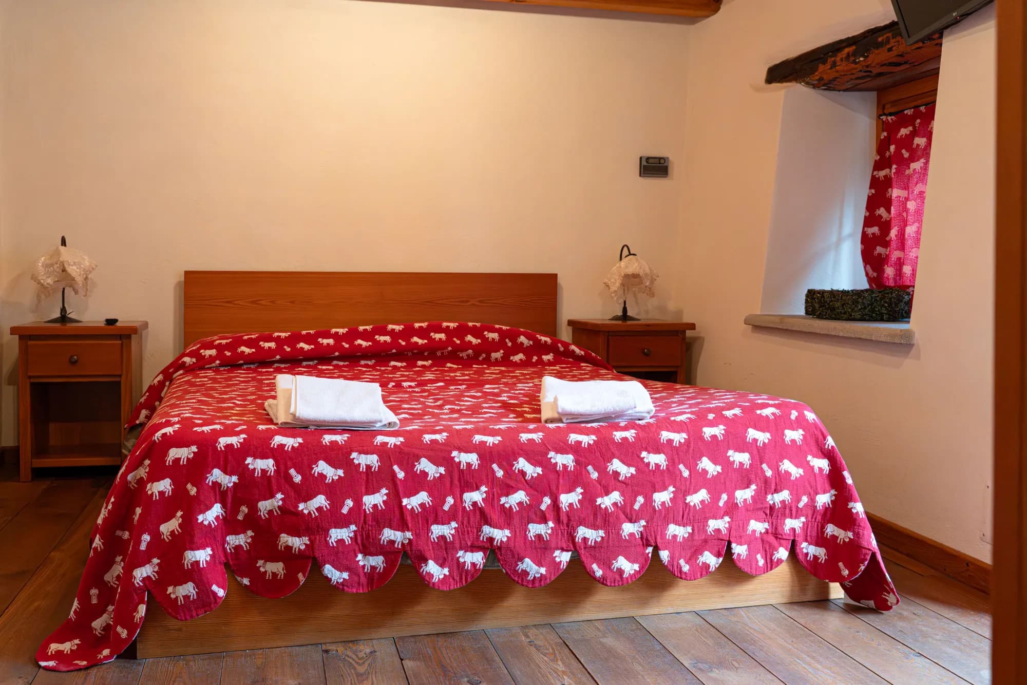 Camera Bucaneve: letto matrimoniale con copriletto rosso a cavallini bianchi, tende in tinta alla finestra incassata nel muro in pietra, pavimento e trave in larice