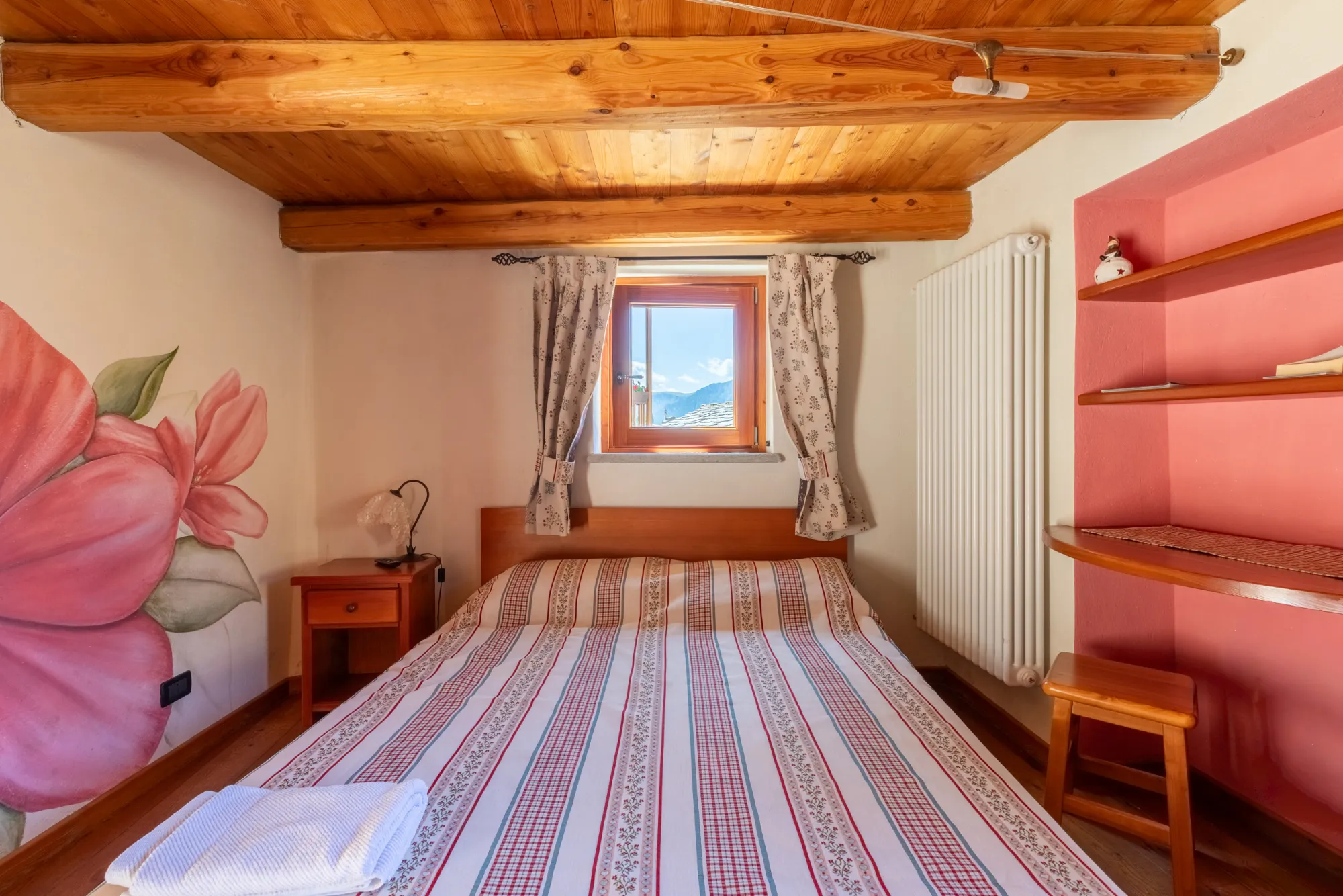 Camera Rododendro: letto matrimoniale sotto il soffitto a travi di larice, finestra aperta sulle montagne e grande rododendro dipinto sulla parete accanto alla nicchia rosa